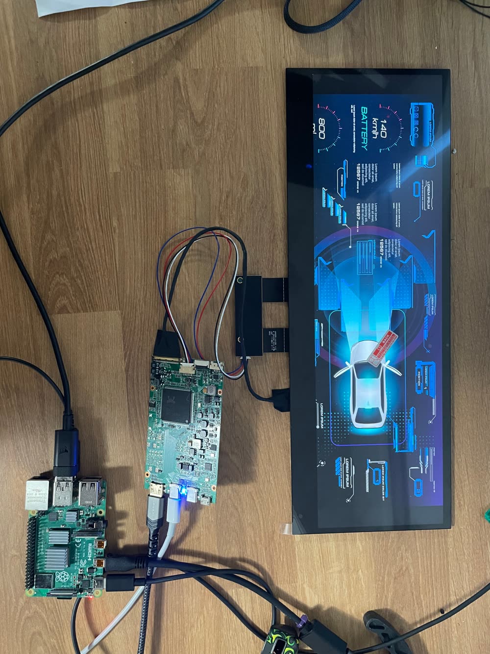 On a wooden floor, a Raspberry Pi and a display controller board wired to an ultrawide screen showing a placeholder car dashboard