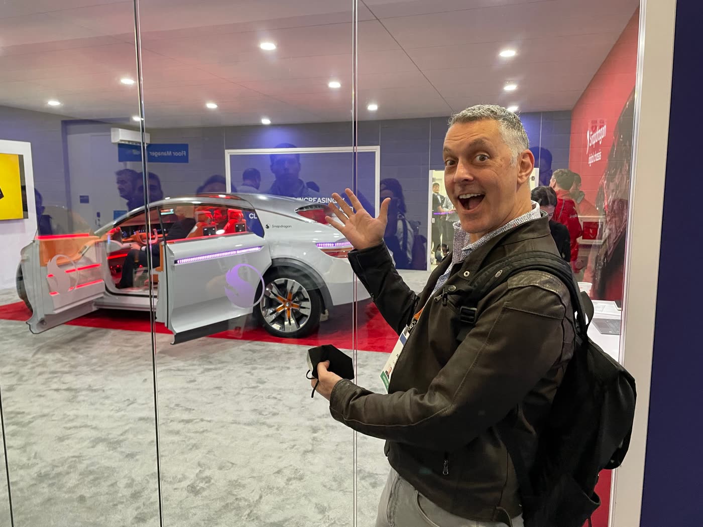 Joe grinning and gesturing at the Snapdragon concept car, doors open and cabin lit red and purple, behind glass at the Qualcomm booth