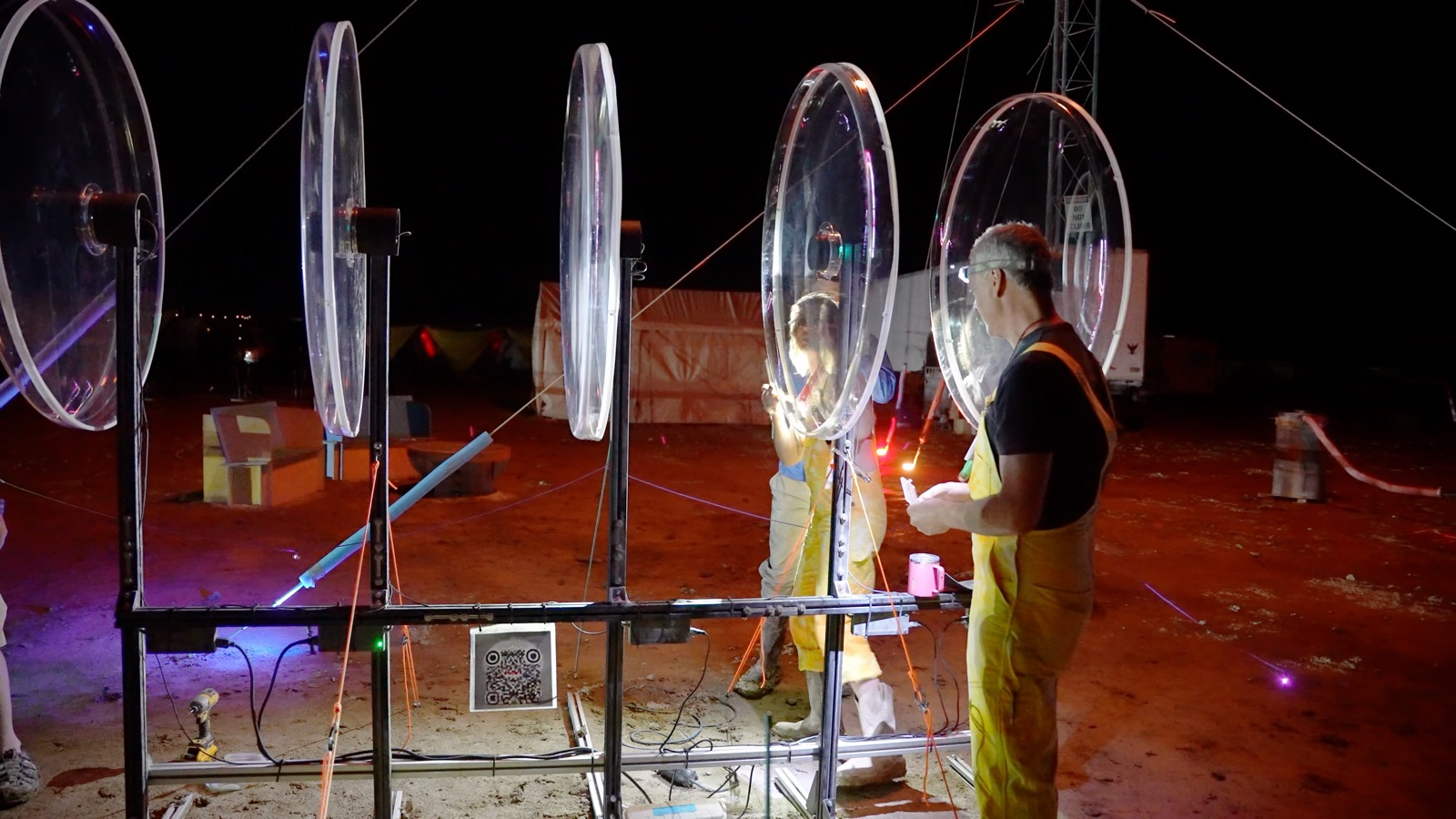 At night on the playa, two people in work clothes tune five large clear circular blades mounted on a steel frame
