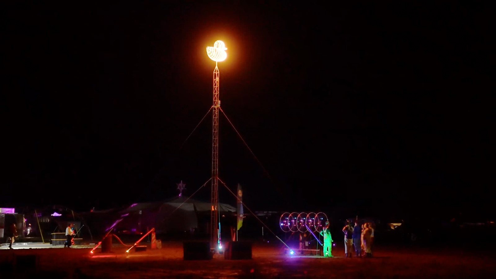 The Blades lit up purple at the edge of a dark camp on the playa, beside a tall lit tower and a few people