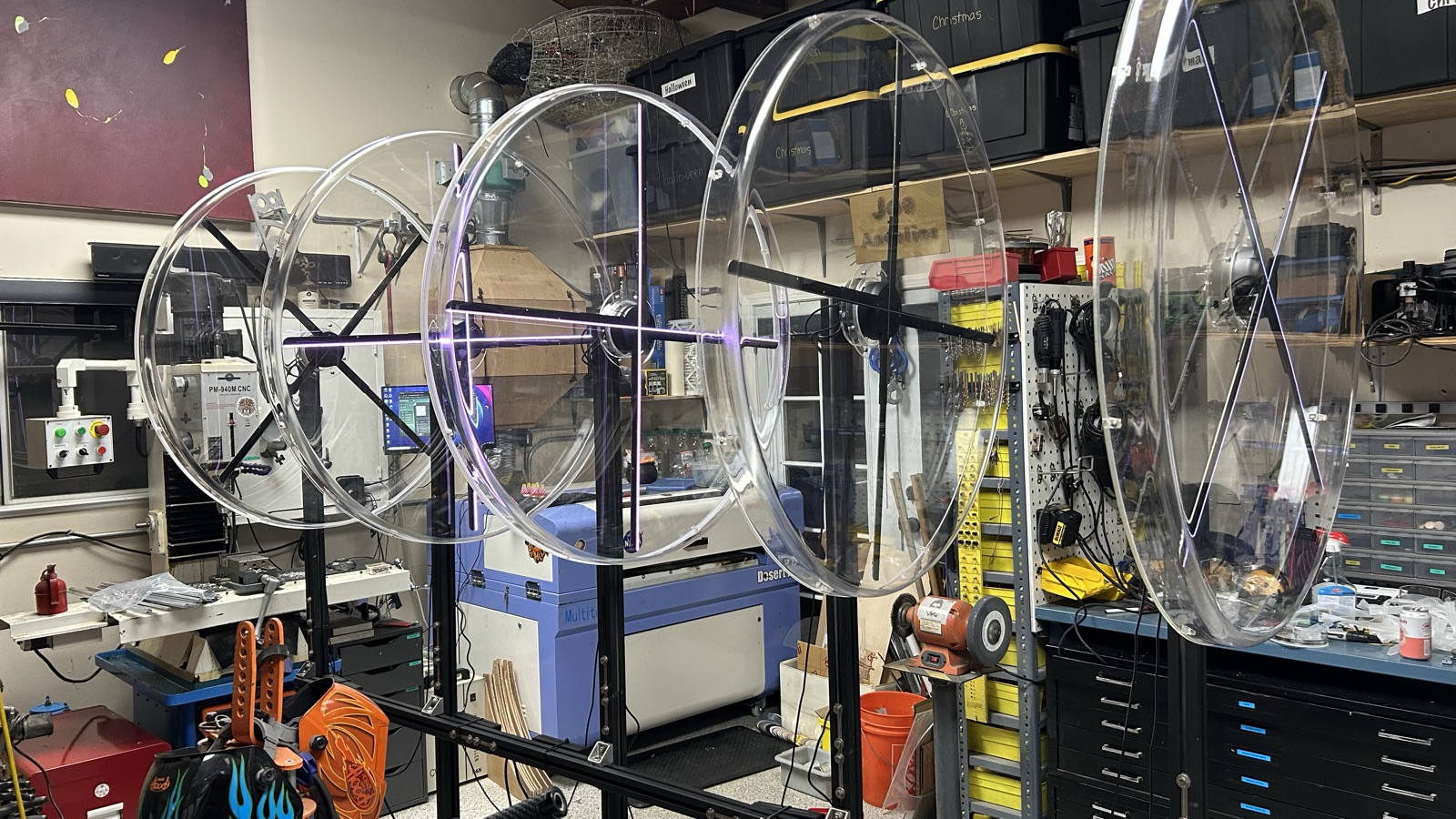 In a workshop, five large clear circular blades stand in a row on a steel frame among shelves, tools, and a laser cutter