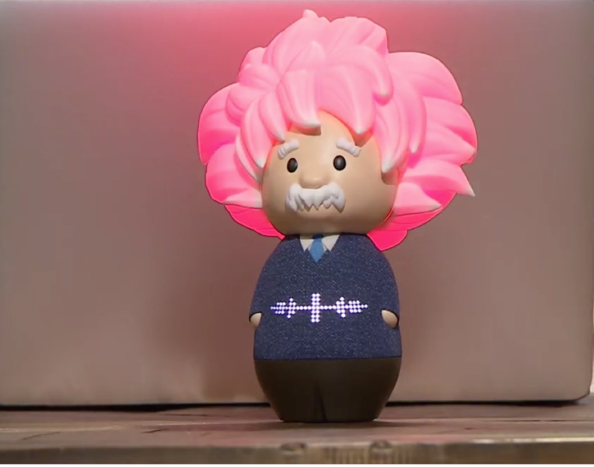 The Einstein smart speaker on a table: a figure with glowing pink hair, a white mustache, and a navy sweater with a lit-up voice waveform across the chest