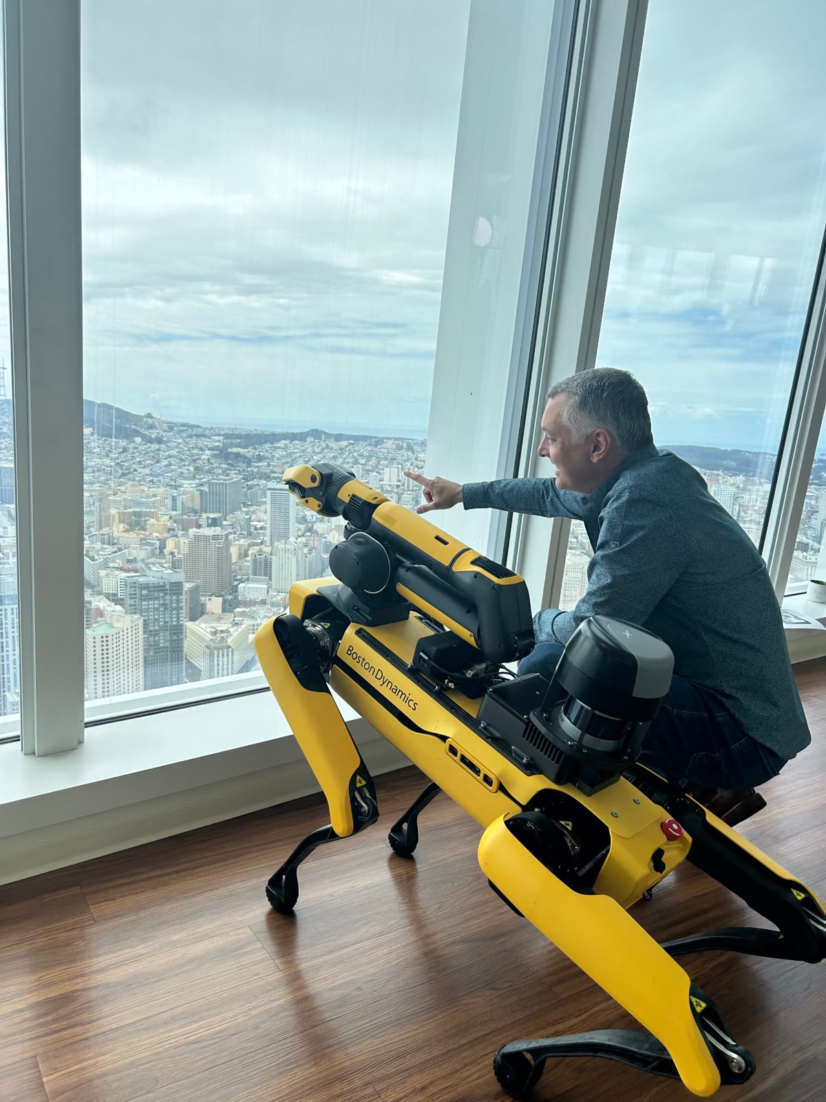 Joe Andolina kneeling beside a Boston Dynamics Spot robot at a window high above San Francisco, pointing out at the city