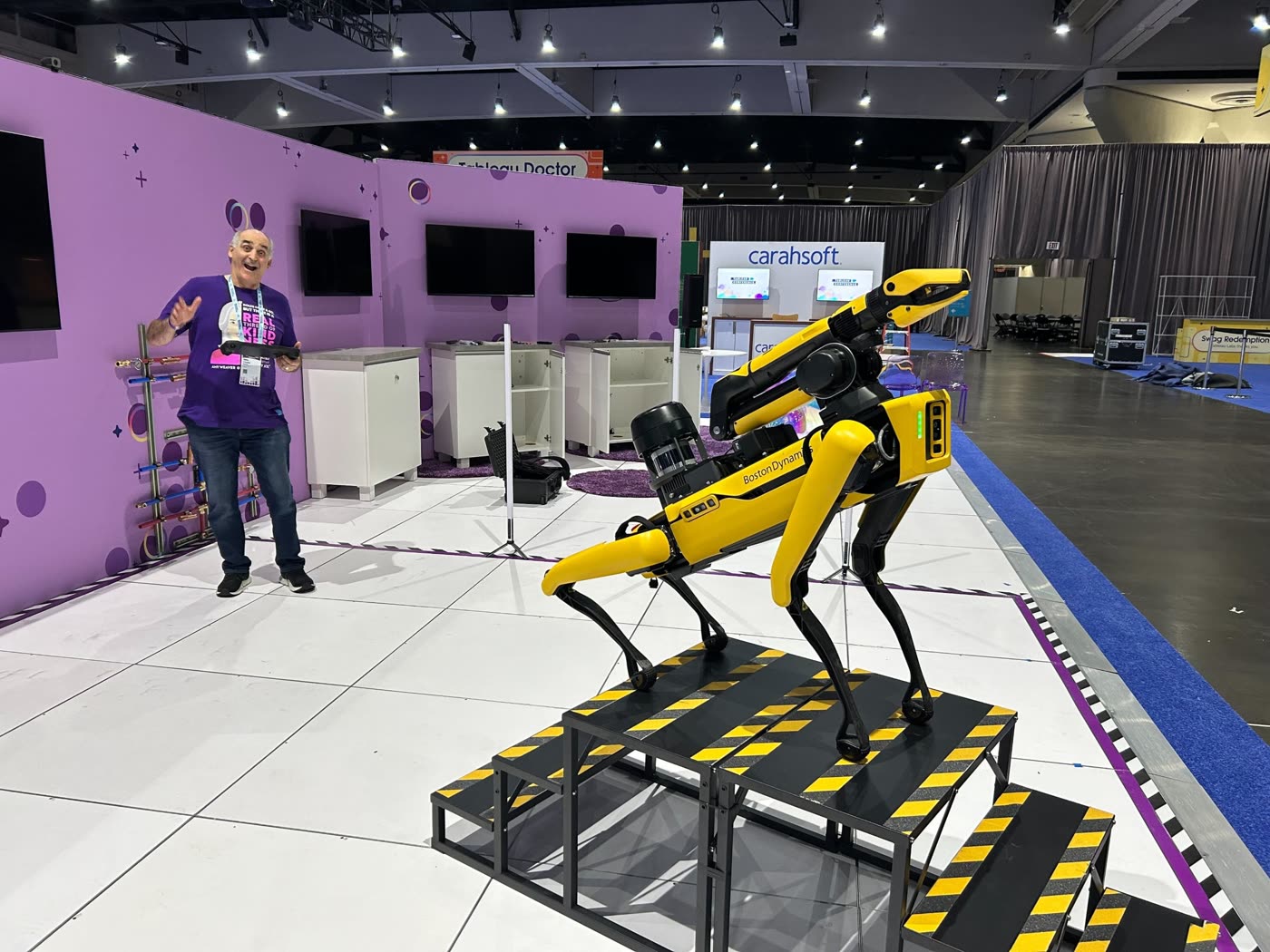 Spot standing on yellow-and-black hazard-striped stairs at a purple show booth, while a teammate holding Spot's controller laughs in the background